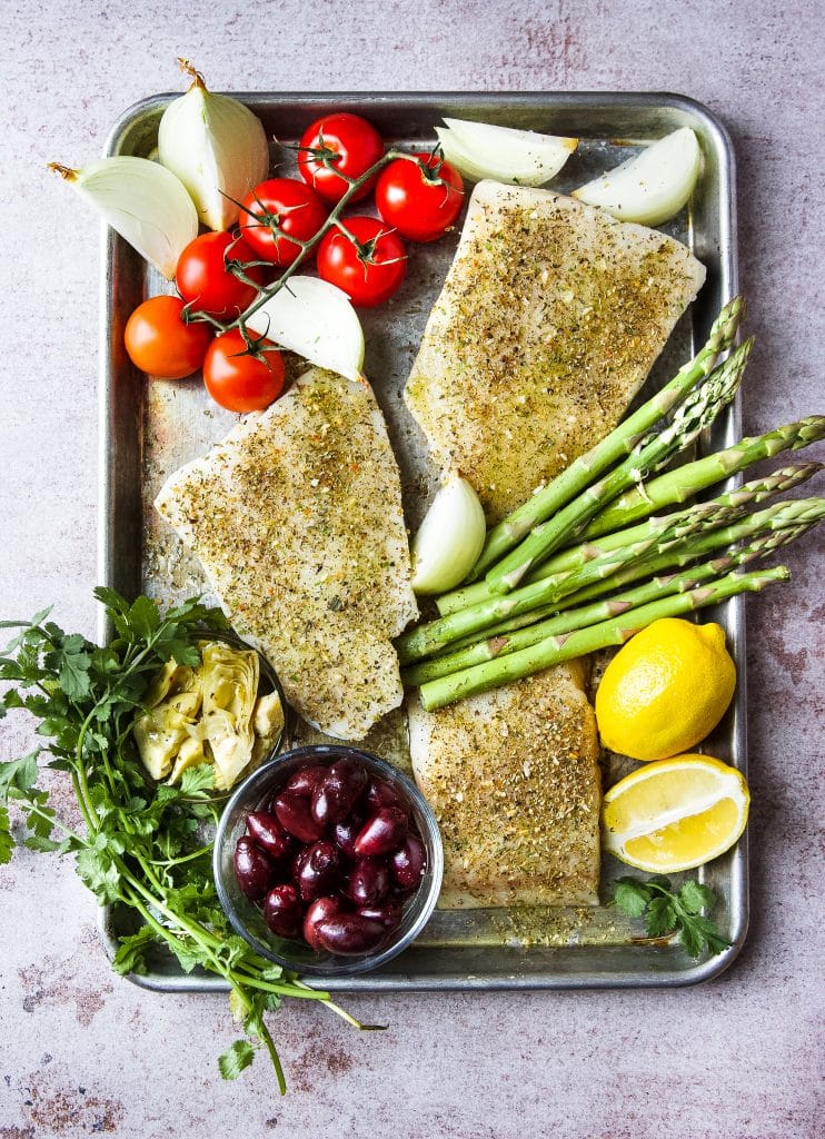Three cod filets seasoned in sheet pan. A selection of vegetables such as asparagus, tomatoes, onions, olives, cilantro and lemon in sheet pan ready tote cooked. 