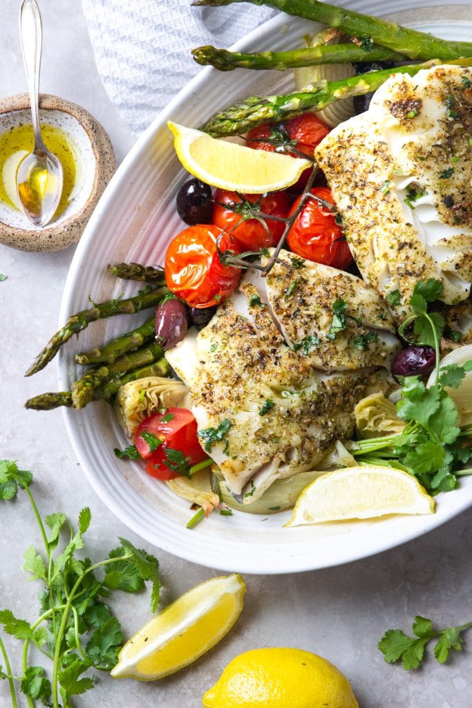 Herb seasoned cod filets in platter with tomatoes, asparagus, olives, herbs and lemon wedges. 