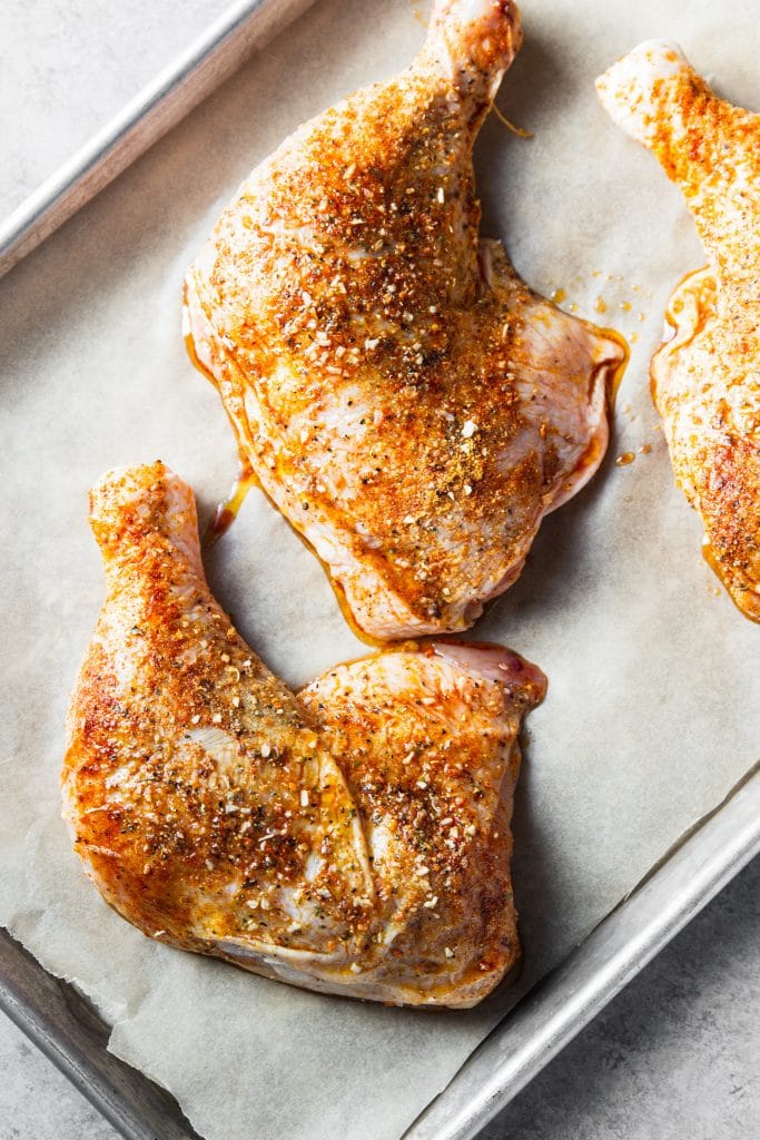 Raw chicken coated with olive oil and spice rub on a sheet pan lined with parchment paper.