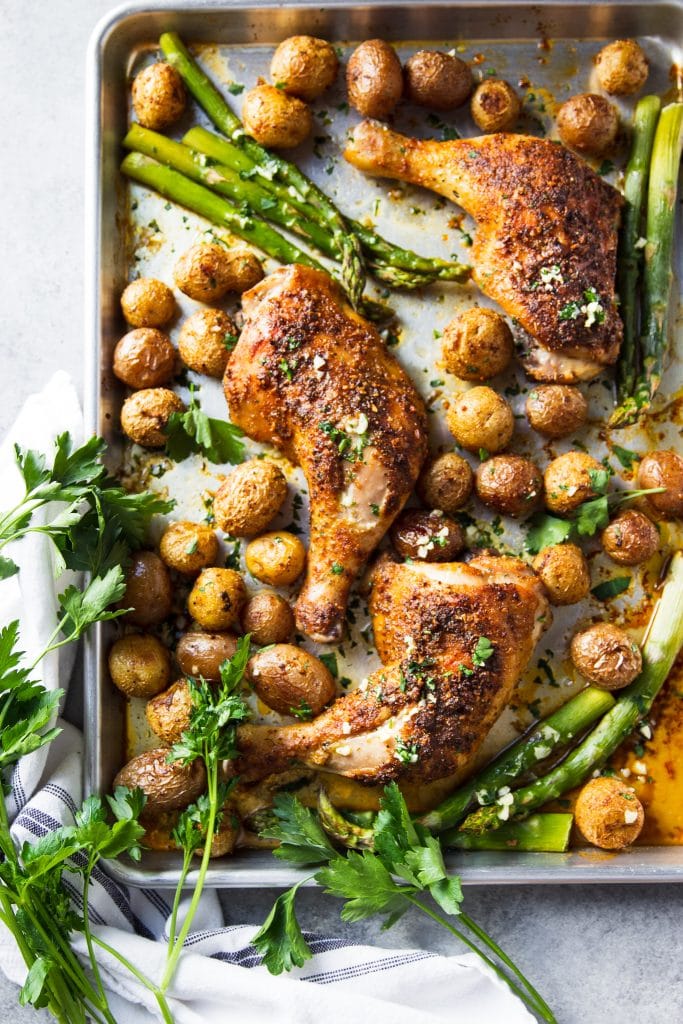 Roasted chicken, baby potatoes, parsley and asparagus, on a baking sheet lined with parchment paper. 
