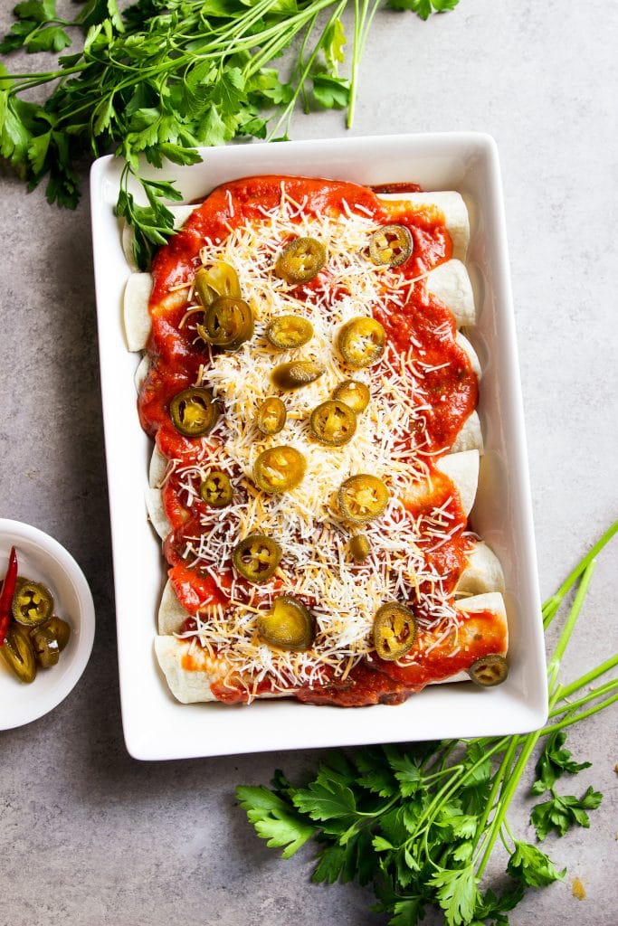 beef enchiladas topped with tomato sauce, cheese, and jalapeno slices in a white baking dish.