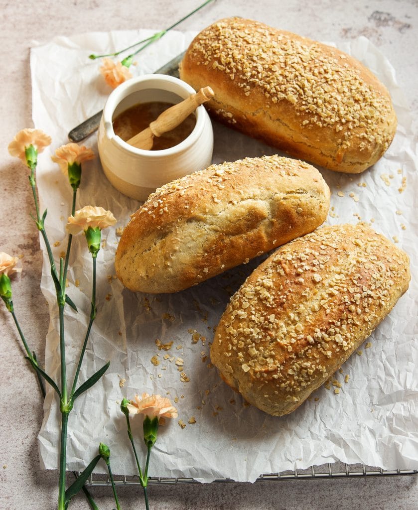 three loaves of honey oat bread on a cooling rack lined with parchment paper. A small pot with honey. Pink flowers on the table