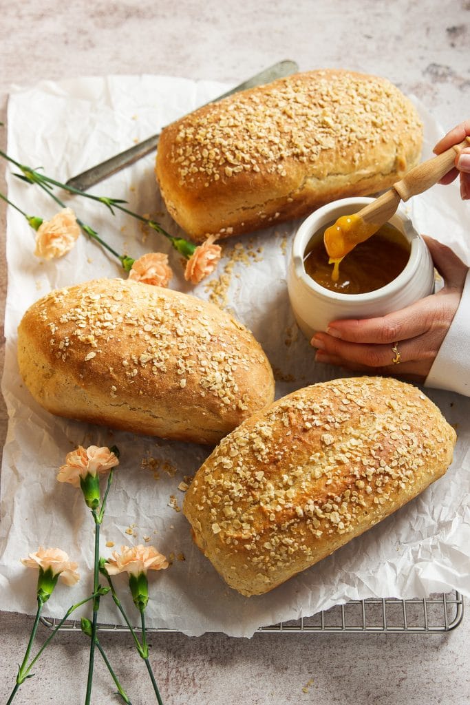 three loaves of honey oat bread on a cooling rack lined with parchment paper. A hand serving a small pot with honey. Pink flowers on the table