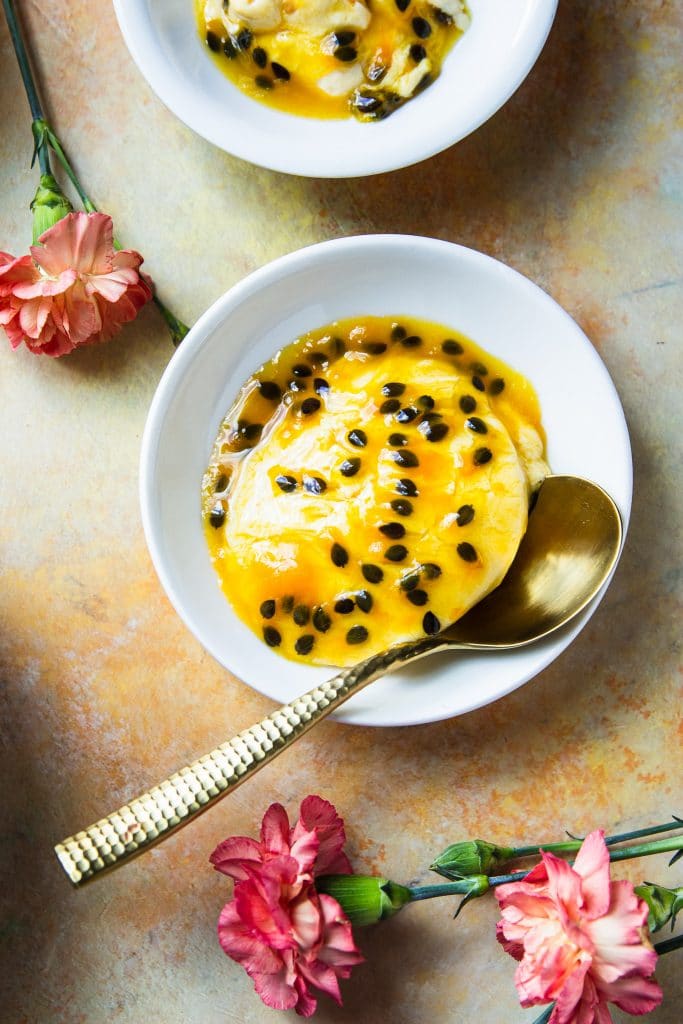 a small white plate with passion fruit mousse and a spoon. There are pink flowers on the table.