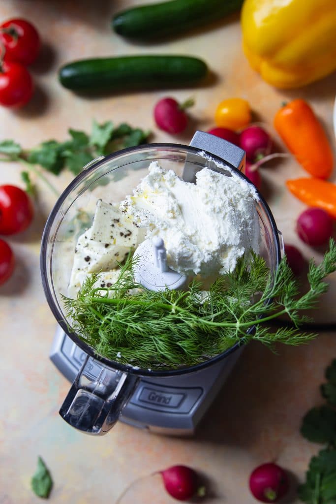 feta cheese, dill, and cream cheese in a food processor. Fresh veggies around the table. 