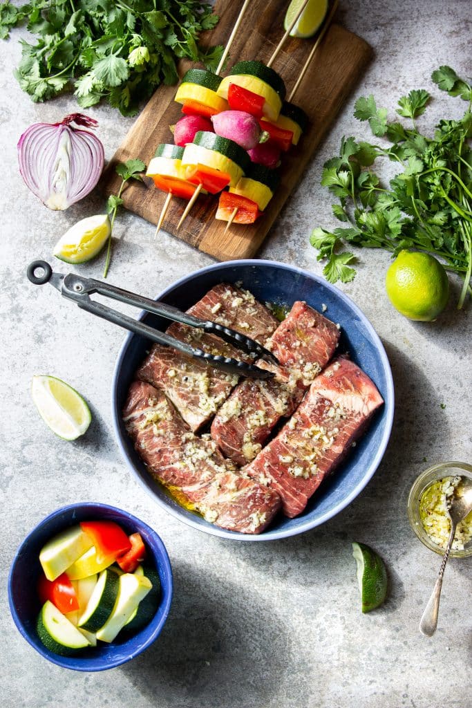 Seasoned flat iron steaks for churrasco. Fresh veggies in a kabob. Herbs and lime slices on the table. 