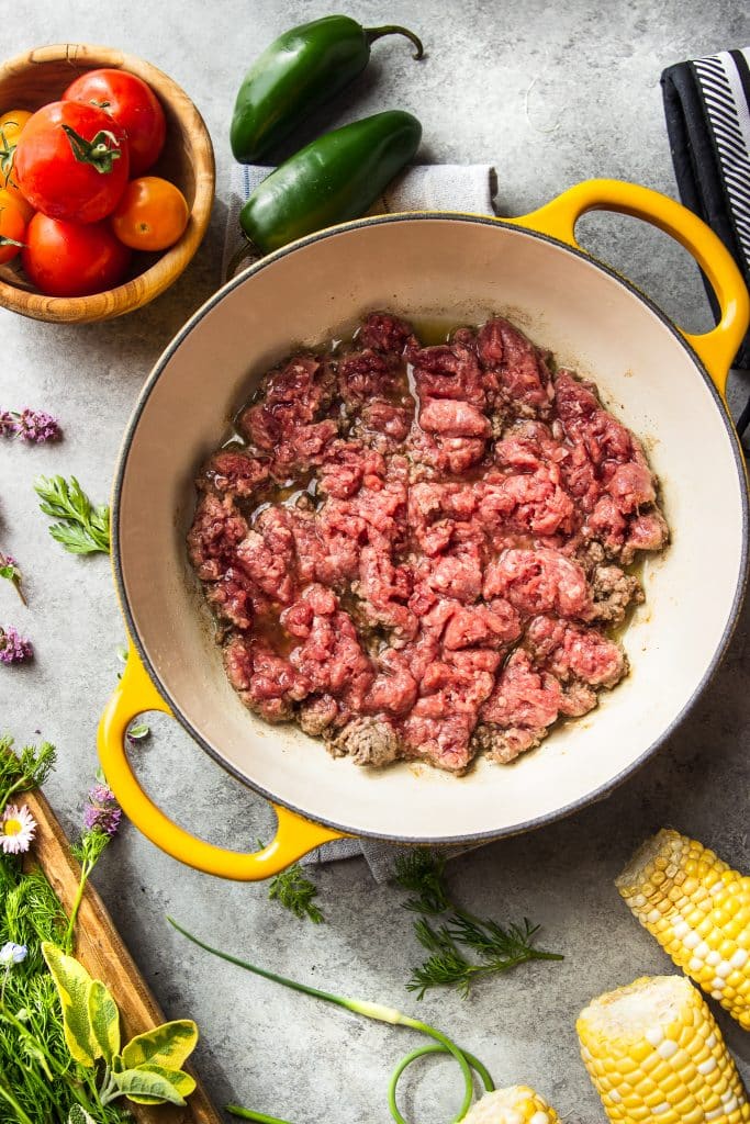 Ground beef being cooked in skillet with oil. Fresh herbs and corn on the cob, on the table. 