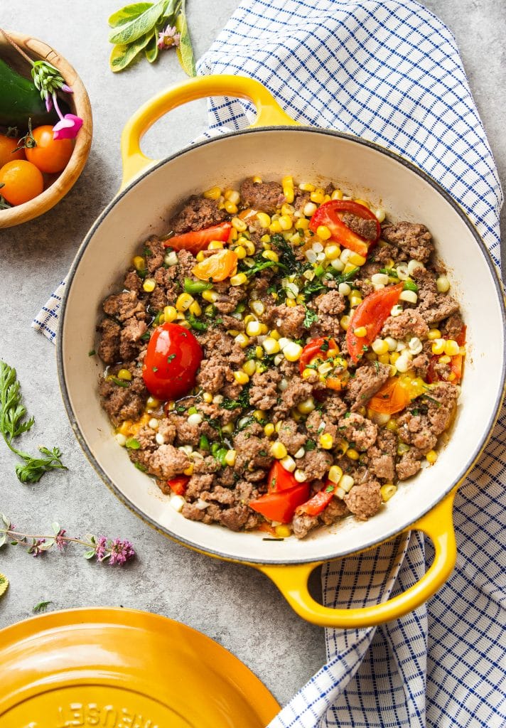 Ground beef in skillet with corn, tomatoes and fresh herbs. A table cloth under the skillet and a small wooden bowl with tomatoes on the table. 