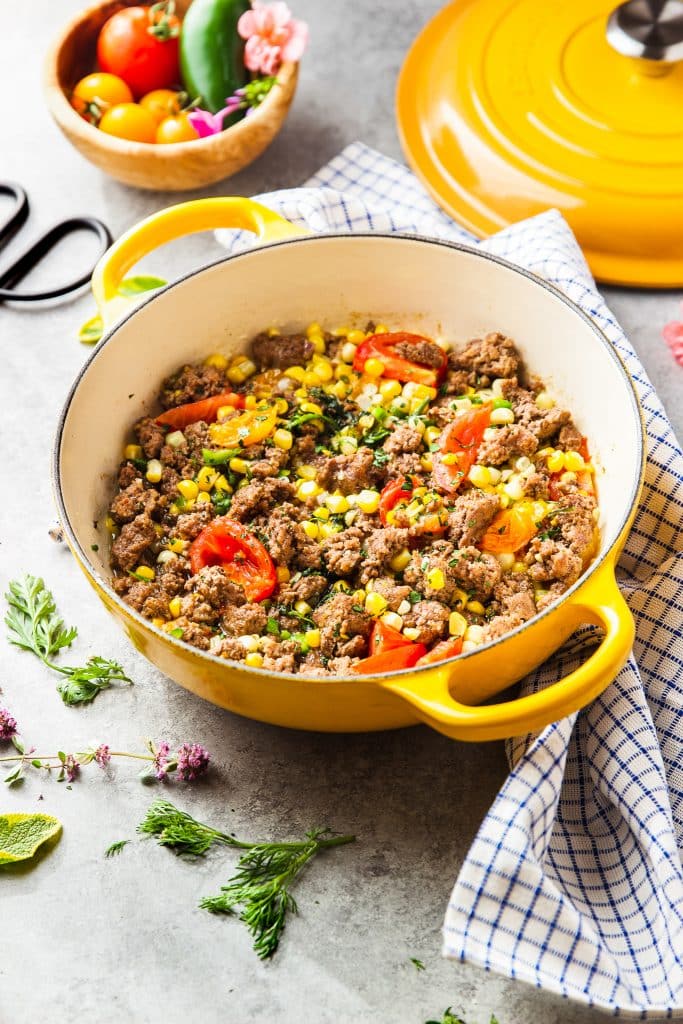 Ground beef, corn and tomato skillet. Fresh herbs and a table cloth around it.