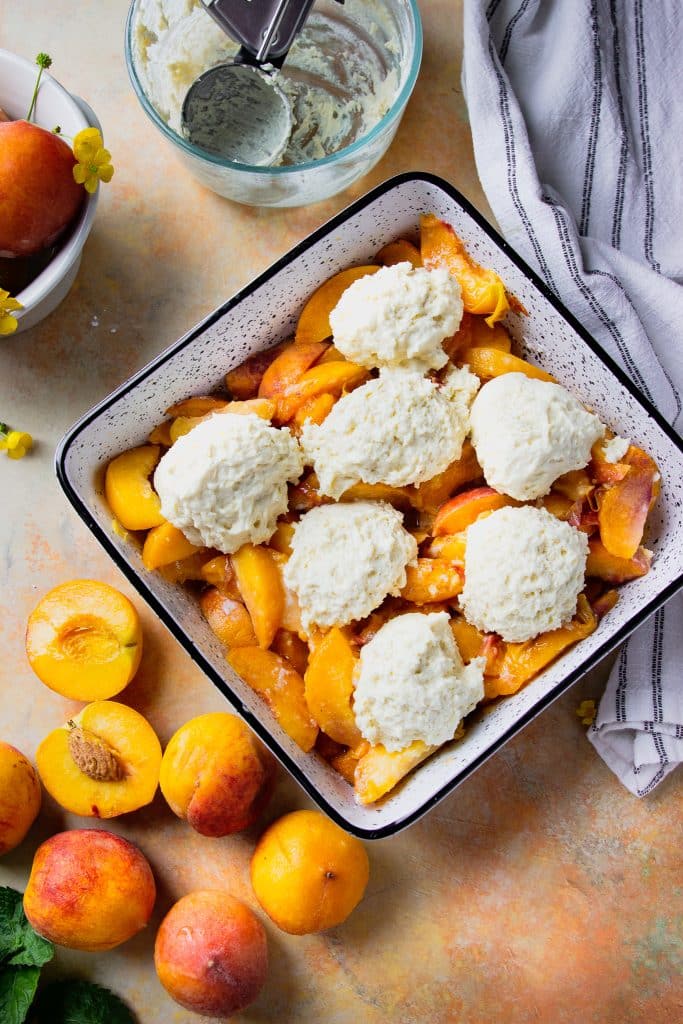 Peach slices in baking dish topped with biscuit dough. Extra peaches on the table, and a table cloth.