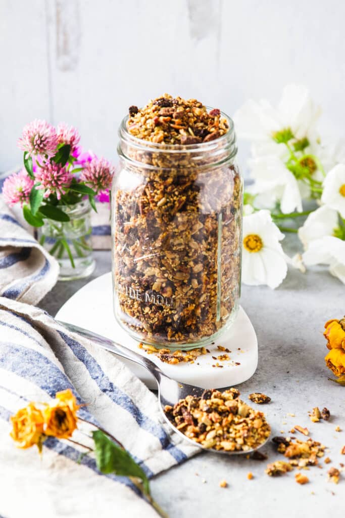 A jar of grain free granola. some flowers on the background