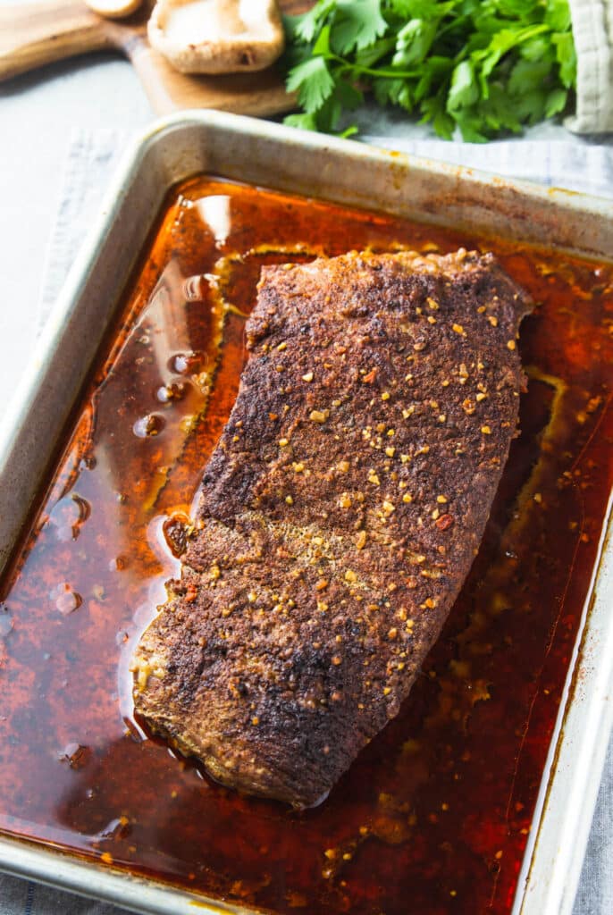 Beef brisket out of the oven, sitting on the meat juices in a sheet pan.