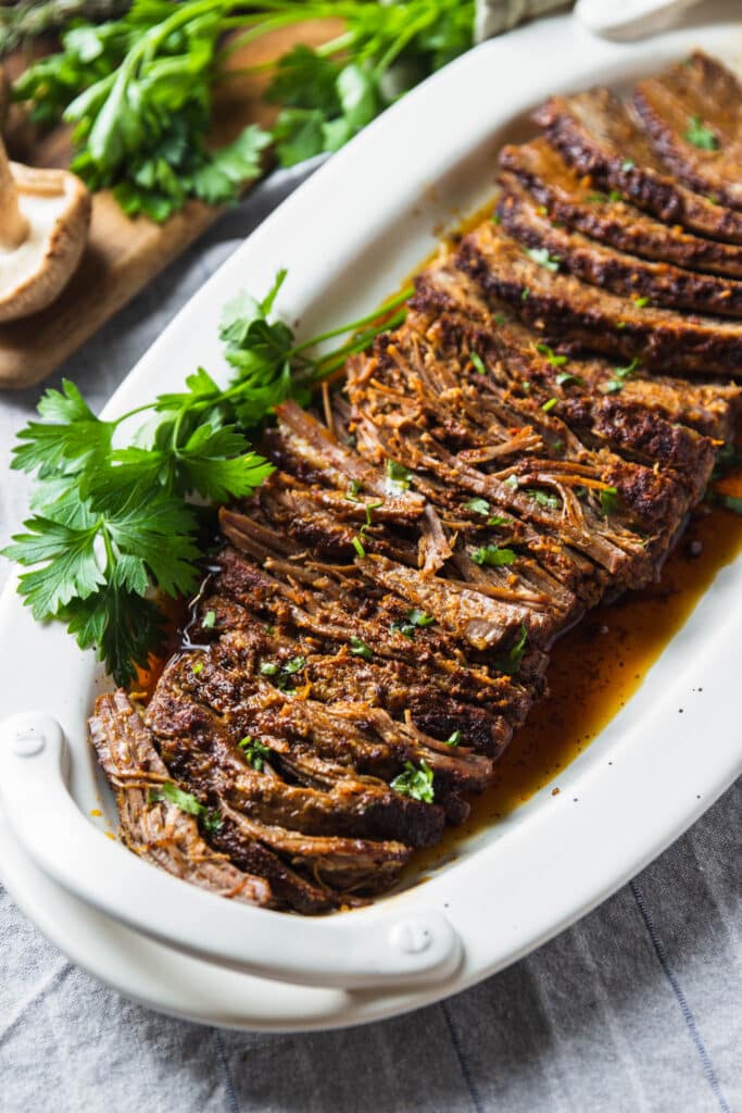 A serving platter with slow cooked oven roasted beef brisket sliced. Fresh herbs on the table.