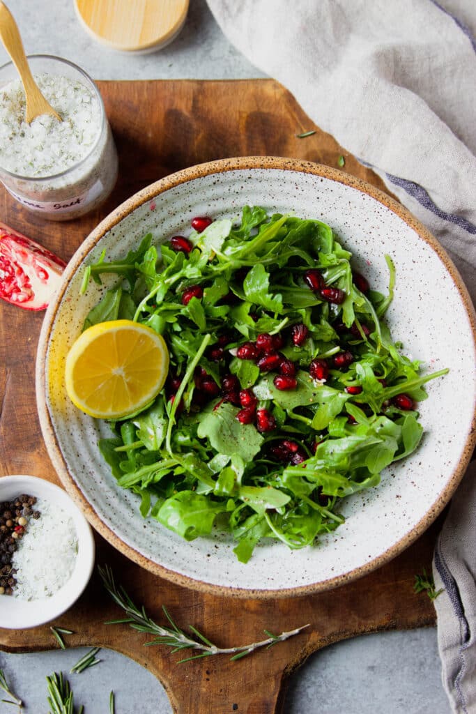 arugula salad with pomegranate seeds, lemon slice and fresh herbs, salt and pepper
