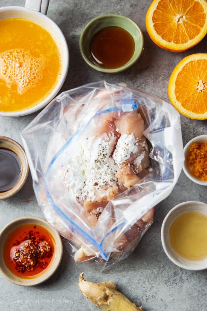 ingredients for air fryer orange chicken. Cubed chicken in ziplock with cornstarch, salt and pepper. Other ingredients around the table like, chili, soy, orange juice and zest, lemon, honey and orange slices. 