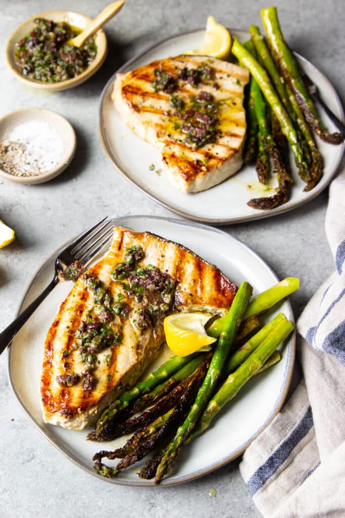 two swordfish steaks topped with olive tapenade and a side of asparagus. Some seasoning in a small bowl and fresh lemon wedges. 