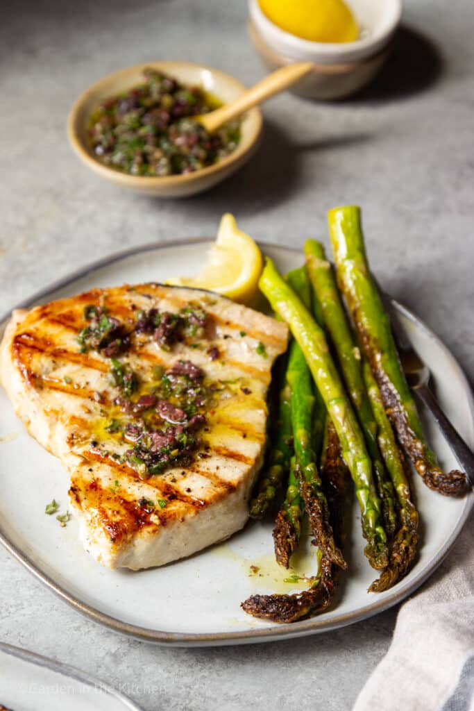 swordfish steak with grill marks, topped with olives and a side of asparagus.