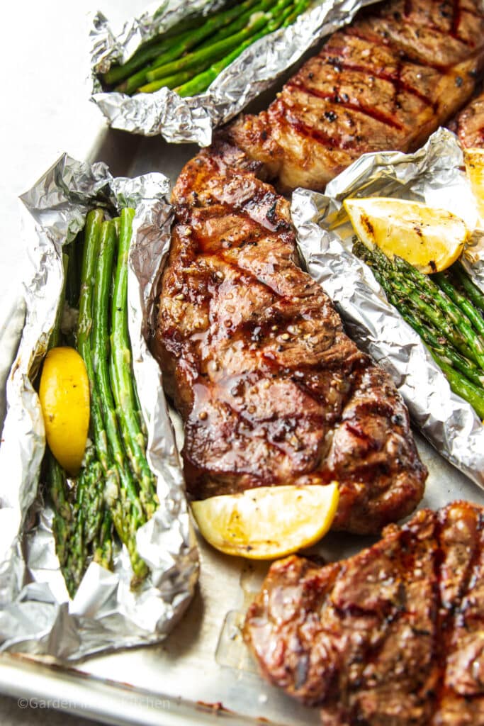 New York strip steak in sheet pan. Grilled asparagus in foil with lemon slices.  