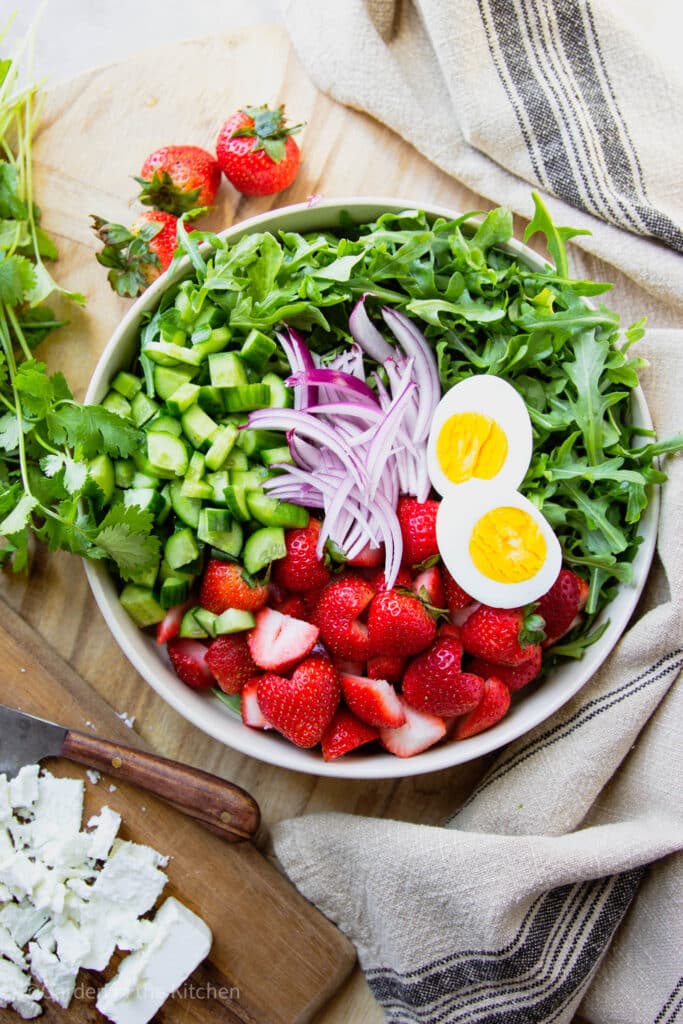 Chopped ingredients for a salad. Cucumbers, sliced strawberries, sliced red onions and hard boiled egg halved in a big bowl with arugula. 