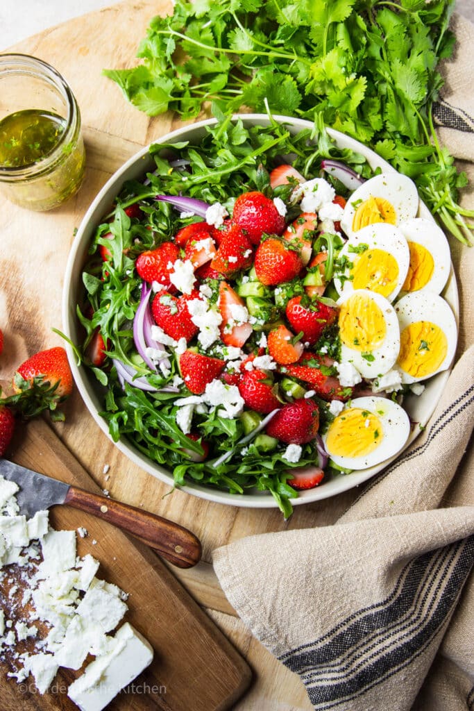 Salad in a bowl with strawberry, arugula, feta, red onions and hard boiled eggs.  Fresh cilantro, a jar of dressing, strawberries and feta cheese on the table. 