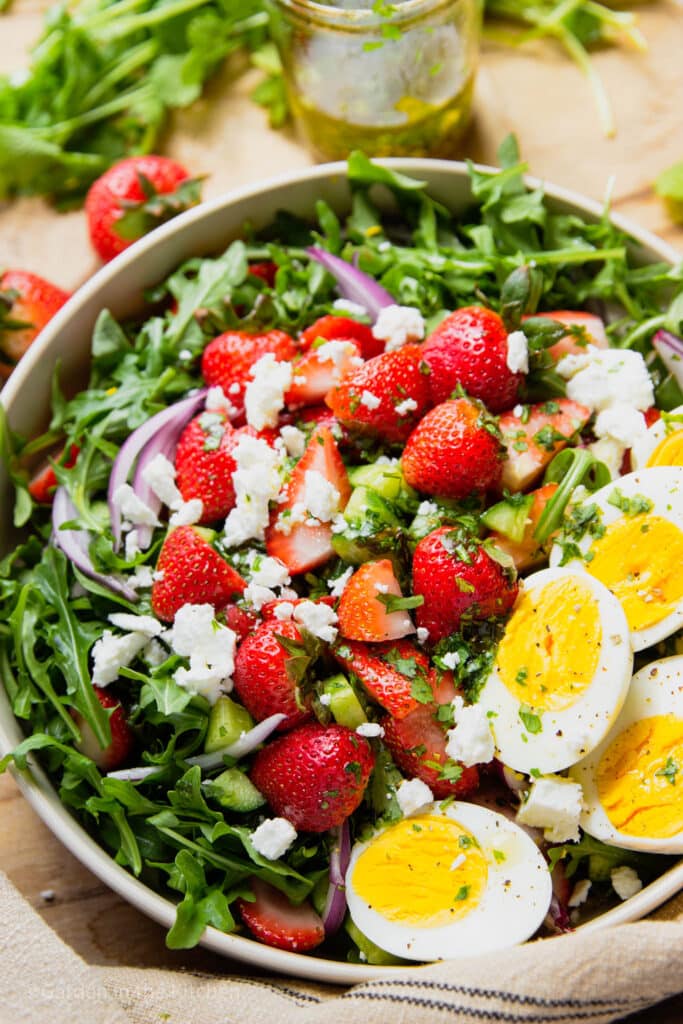 Salad in a bowl with strawberry, arugula, feta, red onions and hard boiled eggs. A jar of salad dressing and fresh strawberries on the table. 