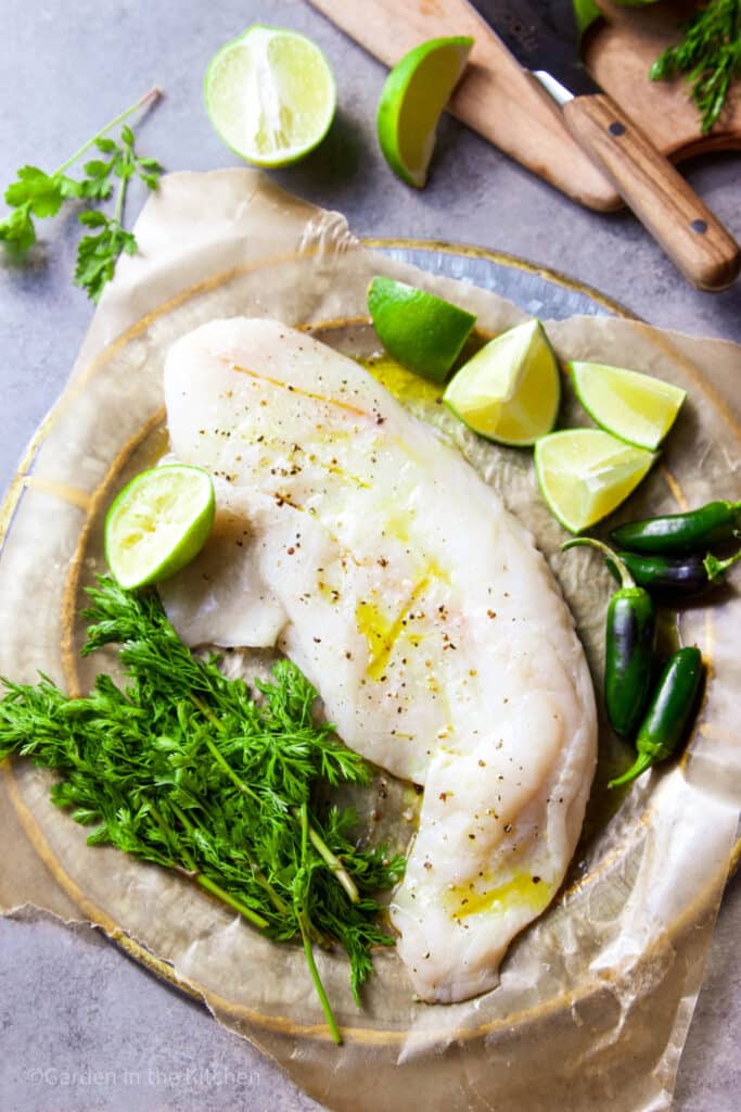 A white fish on a round plate seasoned with salt, pepper and oil. Fresh cilantro, lime and jalapeño on the plate. 