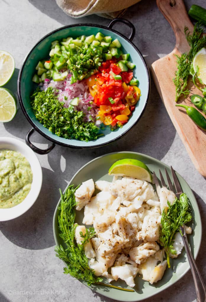 A blue round plate with fresh garden ingredients chopped. A larger green plate with cooked fish, cilantro and lime. Other fresh ingredients on the table. 