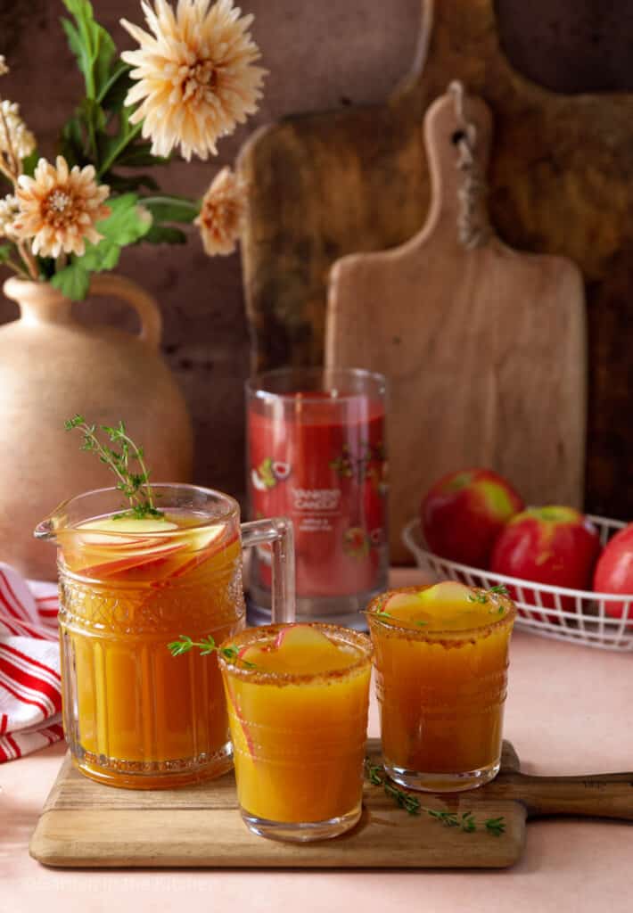 Fall table with apple cider rum punch drink. A candle, a basket of apples flowers and wooden boards in background.