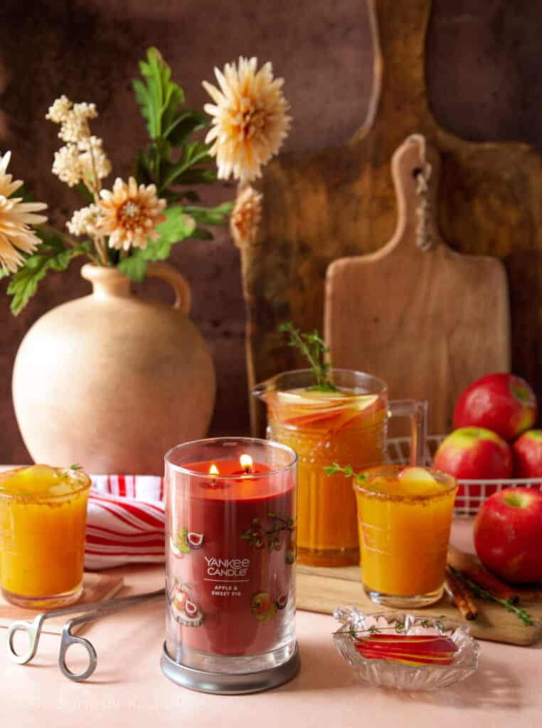 A festive fall table with a red Yankee Candle in the middle. Apple cider rum punch served in glasses and jar. Fresh apples in white wire basket. A red and white stopped kitchen towel. 