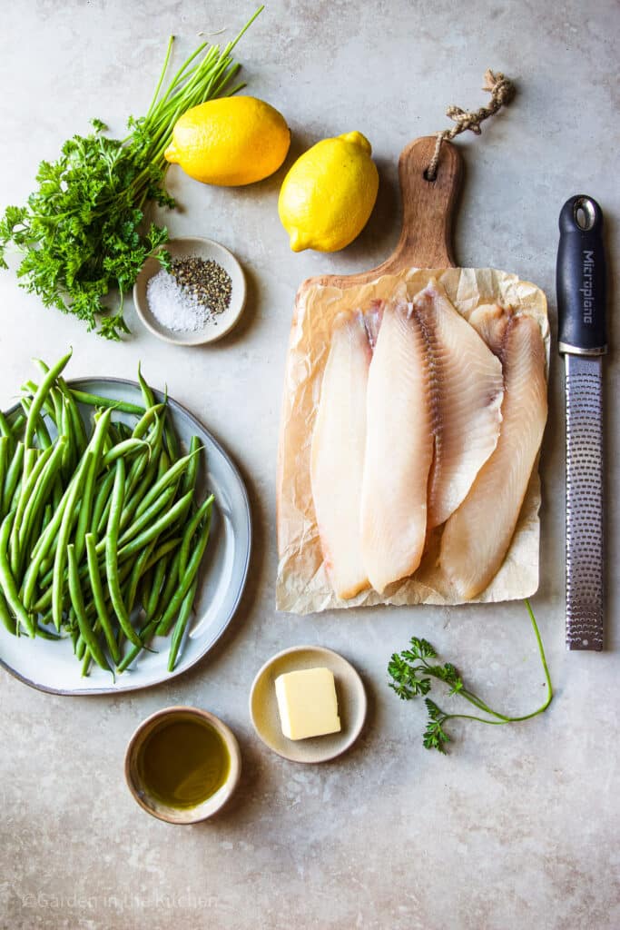 Ingredients for pan seared tilapia. Tilapia filets on a wooden board. Green beans on a gray plate. Small pinching bowls with oil, butter, salt and pepper. Fresh parsley and lemon on the table. 