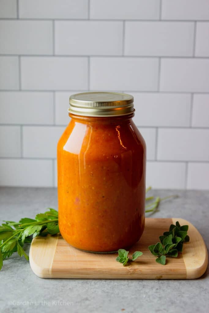 32oz jar of homemade cherry tomato sauce sitting on a wooden cutting board with fresh herbs on the table. 