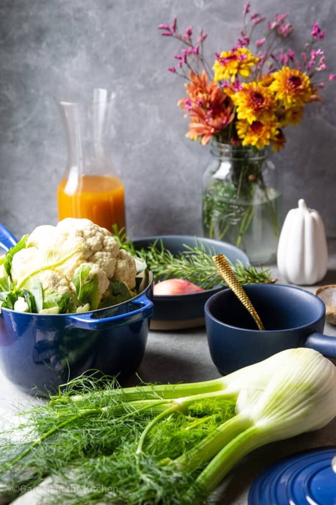 A whole cauliflower inside a blue cast iron pot, uncovered. A flower jar, and white pumpkin and a jar of vegetable broth in background. A whole fennel bulb on the table along with rosemary. An empty blue soup bowl and golden spoon on the table. 
