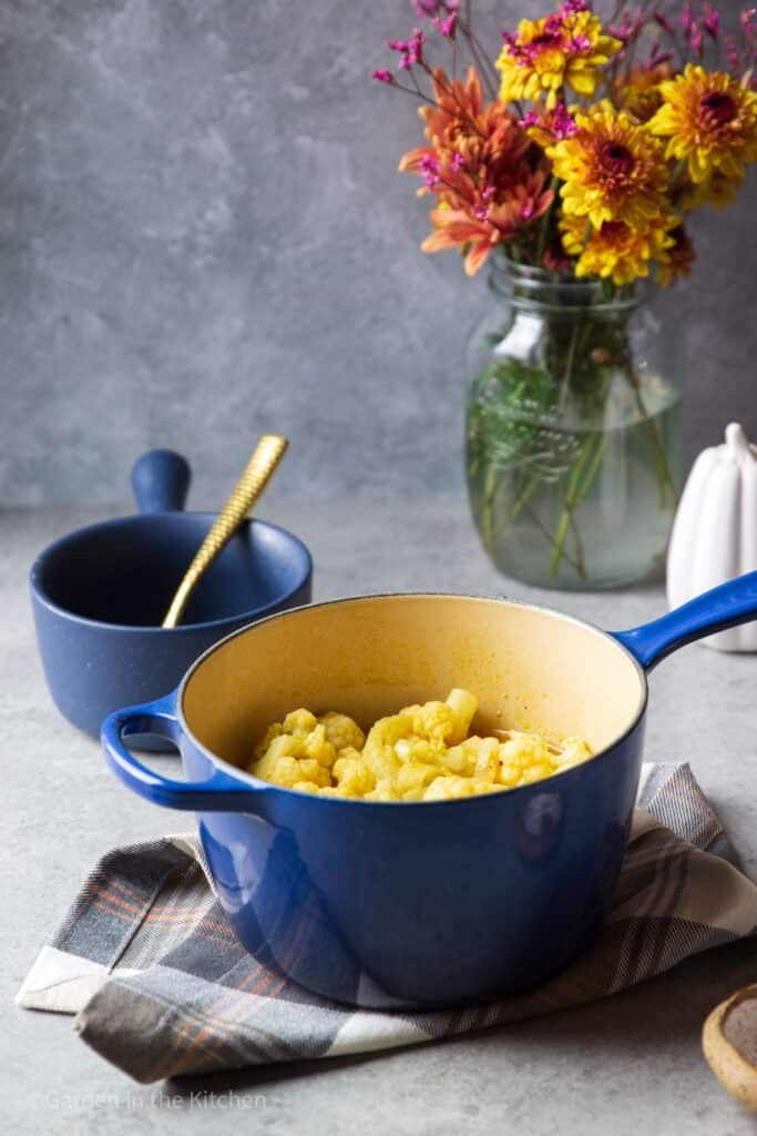 Cauliflower soup contents inside of a blue cast iron pot, sitting on a blue kitchen towel. An empty blue soup bowl with golden spoon in back. 