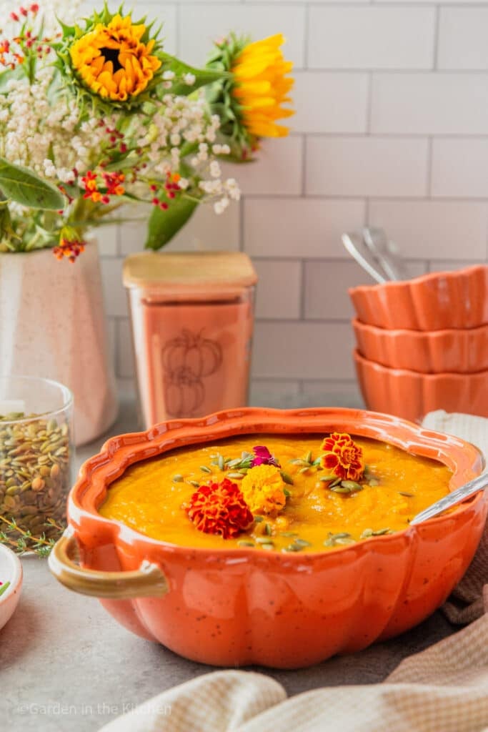 Slow cooker pumpkin soup served in orange pumpkin shaped bowl. Candle and soup bowls in the back. A jar of pumpkin seeds.