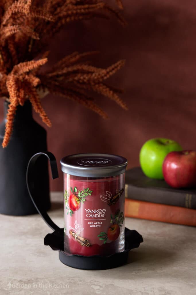 A red Yankee Candle fragrance Red Apple Wreath sitting on a candle holder. In the background two apples on top of two books and a fall arrangement. 