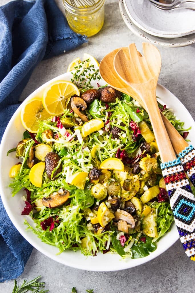 Green salad with roasted squash, Brussel sprouts and mushrooms in a big white bowl. Serving beaded wooden utensils sitting on the side of the bowl.