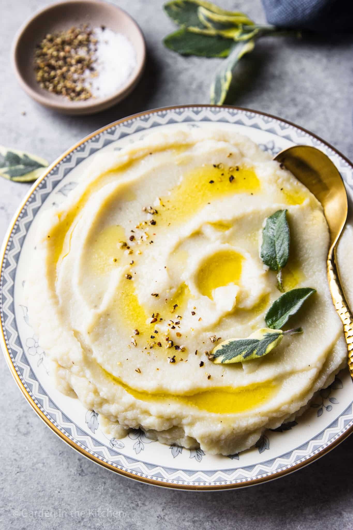 Best cauliflower mashed potato in a round plate with golden and blue details. Cauliflower mash is topped with butter and sage leaves. 