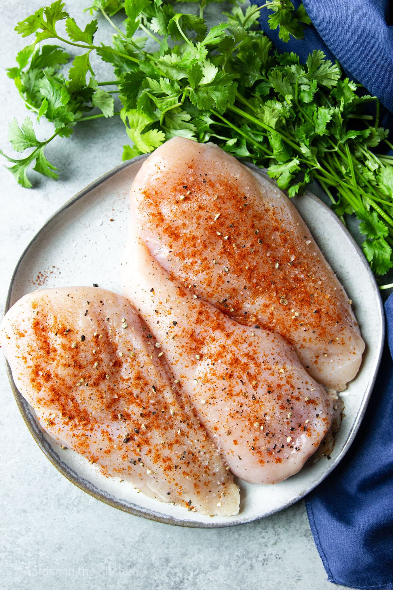 close up on 3 raw chicken breasts seasoned with paprika, salt, and pepper on a white plate.