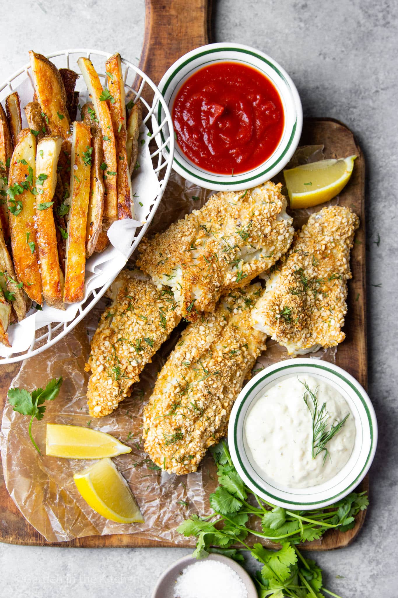 baked panko-coated fish filets on a wood board with small bowls of fries, ketchup, and tartar sauce on the side.