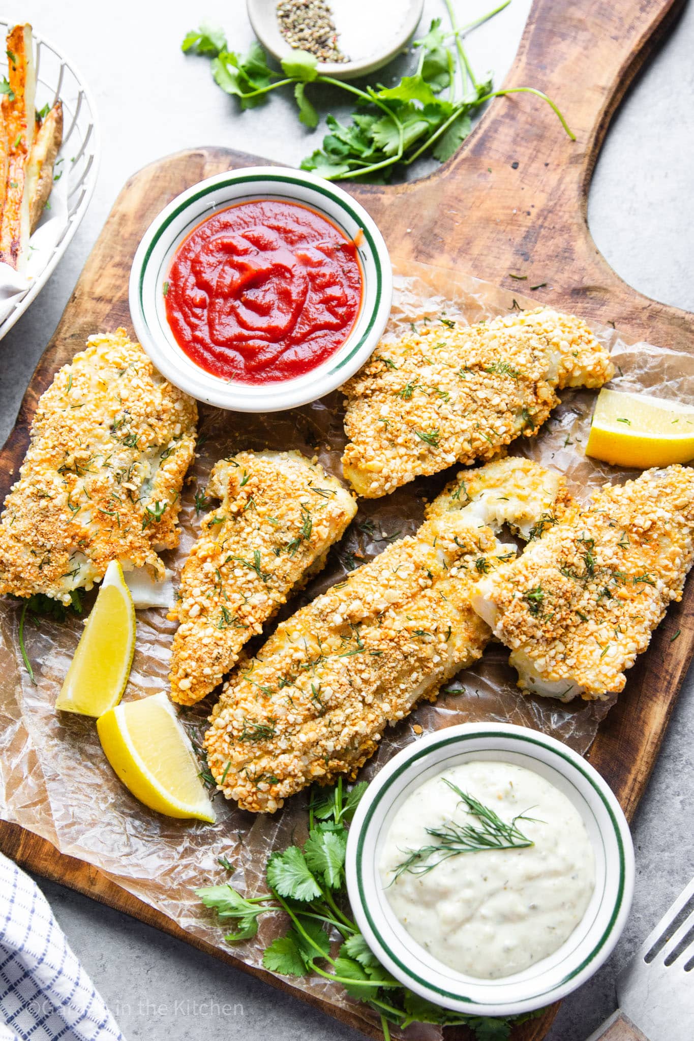 baked panko-coated fish filets on a wood board with small bowls of ketchup and tartar sauce on the side.