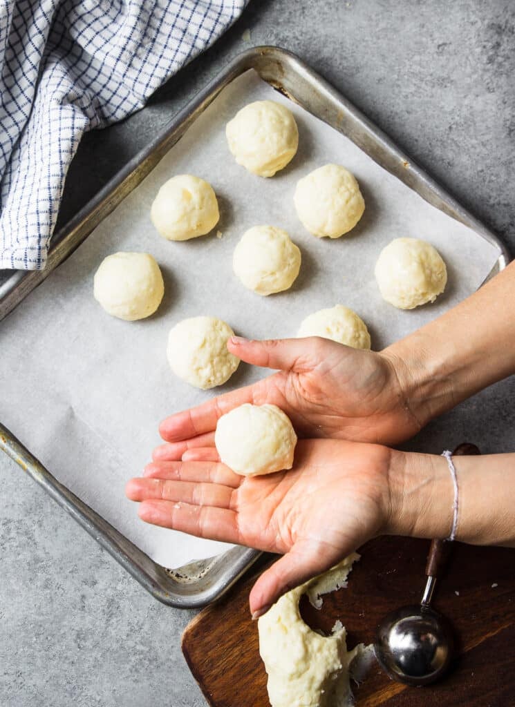 a hand holding a small ball of dough next to rows of dough balls on a parchment lined baking sheet.