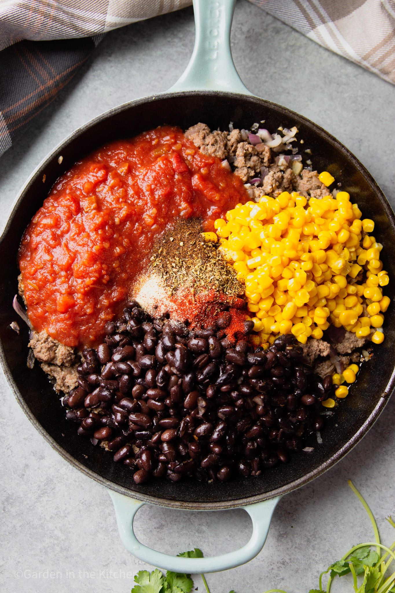 corn, black beans, red salsa, and spices on top of cooked ground beef in a black skillet.