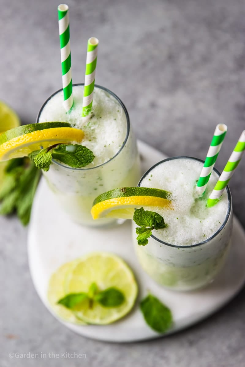 overhead view of 2 glasses of Brazilian lemonade garnished with lime and lemon wedges, mint leaves, and green striped straws.