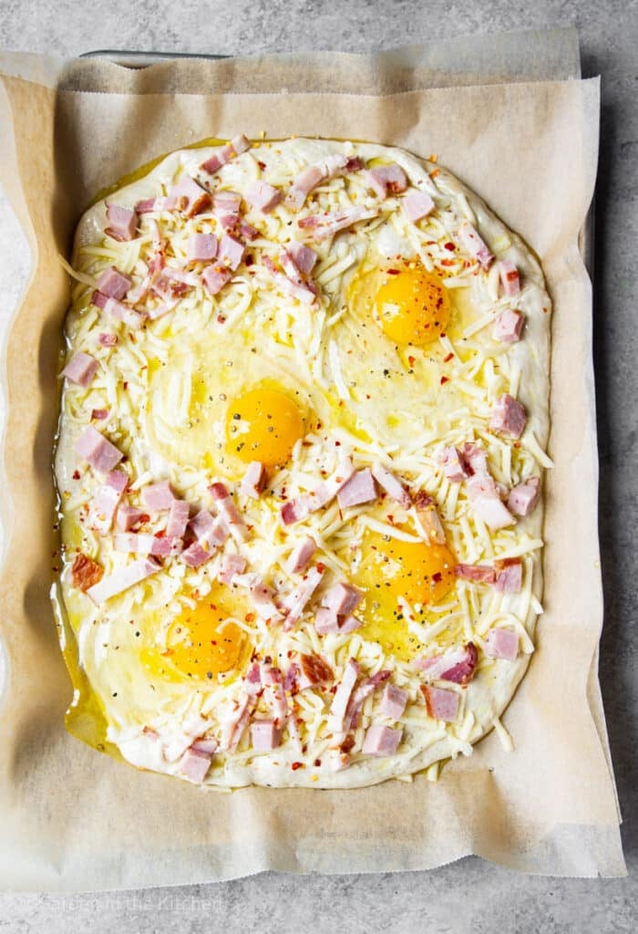 Raw pizza dough with raw eggs, cheese, ham and chili flakes.