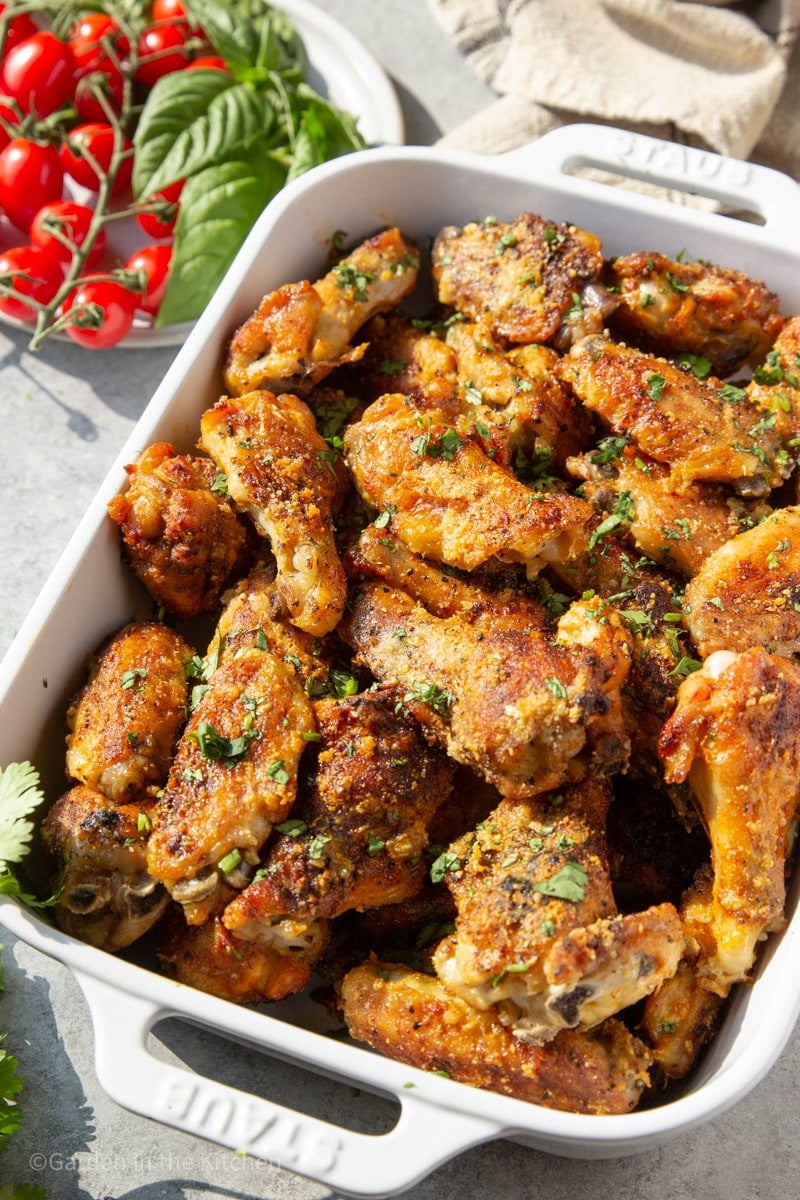 Garlic Parmesan chicken wings in white baking dish, garnished with cilantro. A plate with cherry tomatoes and basil. 