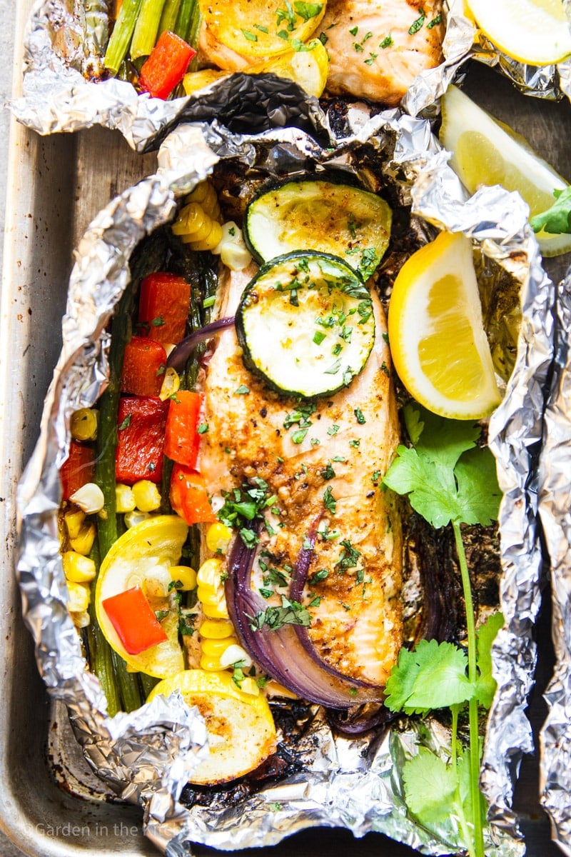 Tin foil packet with grilled salmon, red onion, red bell pepper, corn, zucchini, summer squash, asparagus, cilantro and lemon wedges.