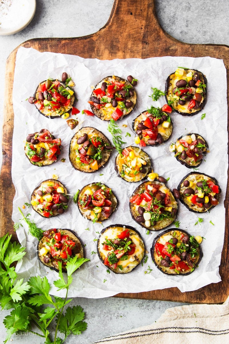 Grilled eggplant tacos; eggplant slices with corn, black beans, cheese, cilantro and tomatoes on parchment paper on a wooden cutting board.