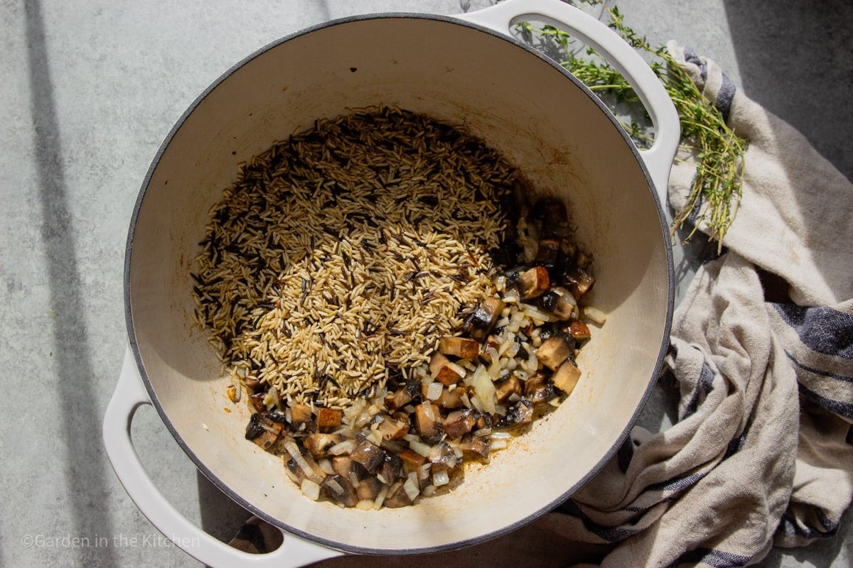 White pot with wild rice, chopped mushrooms, onions, broth, garlic, fresh thyme, and a blue and white dish towel. 