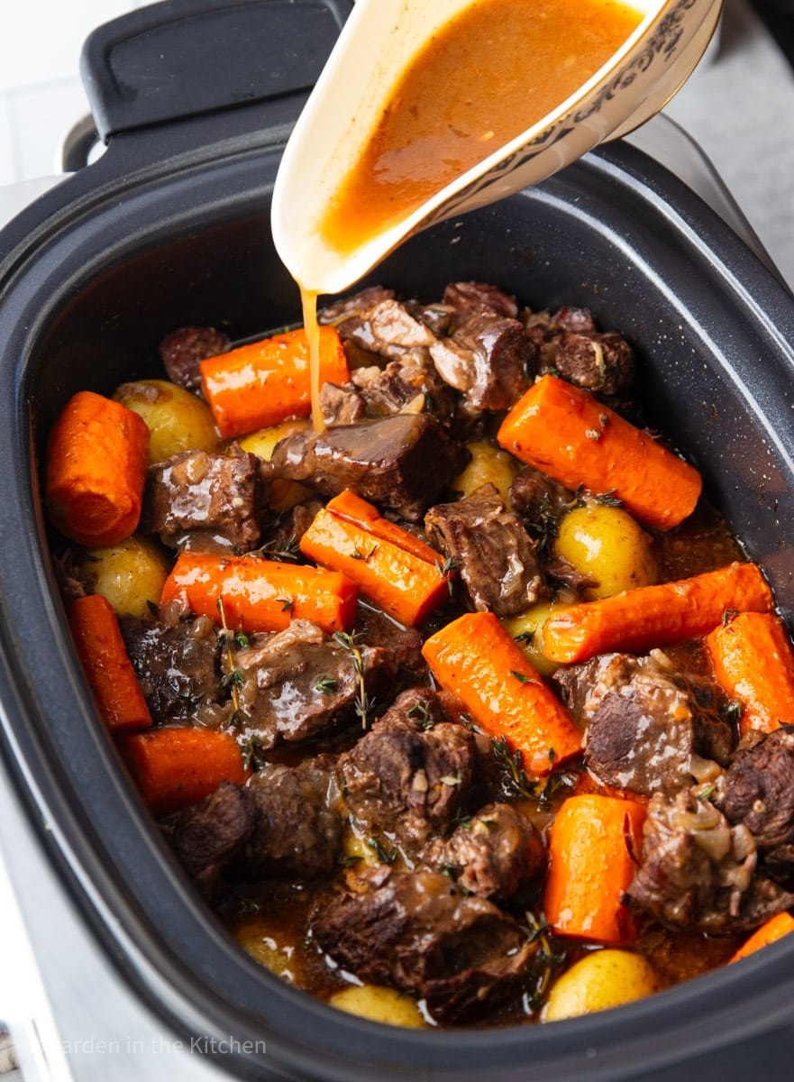 Slow cooker beef stew with chunks of beef, carrots, baby potatoes, thyme in a black slow cooker with broth being poured into the crockpot. 