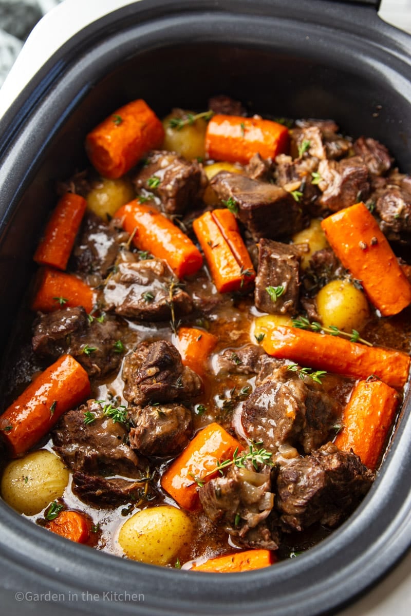 Slow cooker beef stew with chunks of beef, carrots, baby potatoes, thyme in a black slow cooker.